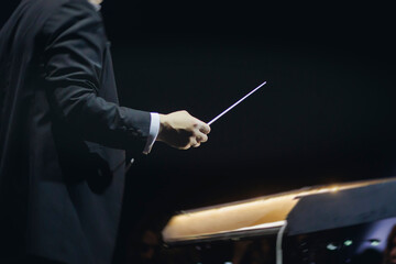Male conductor behind the podium directing orchestra on stage with pupitre, musicians and choir,...
