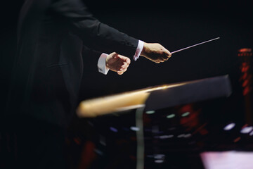 Male conductor behind the podium directing orchestra on stage with pupitre, musicians and choir,...