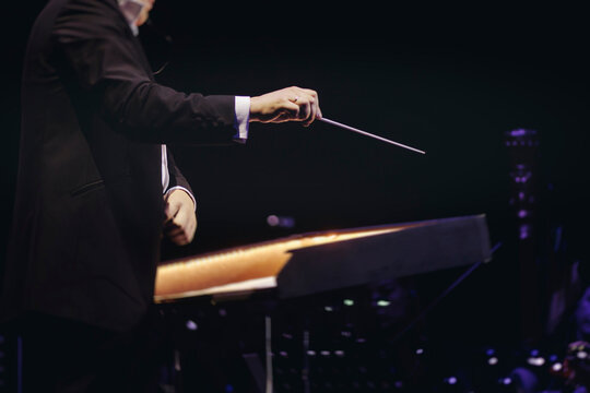 Male conductor behind the podium directing orchestra on stage with pupitre, musicians and choir, the philharmonic venue hall during concert, symphony orchestra director hands conducting waving baton