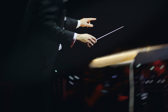 Male conductor behind the podium directing orchestra on stage with pupitre, musicians and choir, the philharmonic venue hall during concert, symphony orchestra director hands conducting waving baton