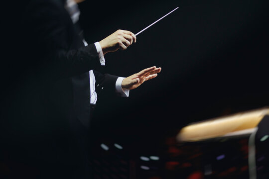 Male conductor behind the podium directing orchestra on stage with pupitre, musicians and choir, the philharmonic venue hall during concert, symphony orchestra director hands conducting waving baton