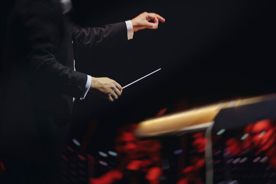 Male conductor behind the podium directing orchestra on stage with pupitre, musicians and choir, the philharmonic venue hall during concert, symphony orchestra director hands conducting waving baton