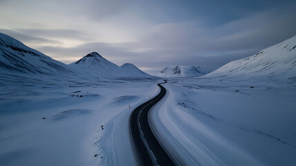 A winding asphalt road cuts through a snowy expanse, nestled between towering snow-covered mountains under a serene sky. The landscape conveys a sense of peace and solitude.