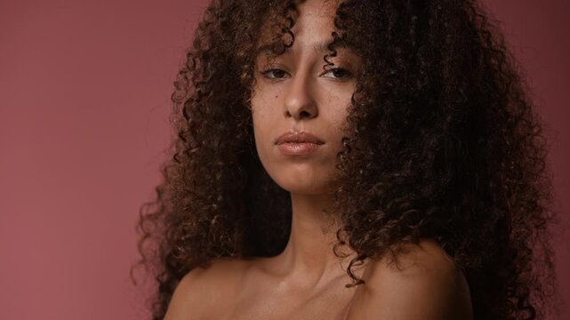 Adorable young adult woman with long dark curly hair, wearing no makeup, looking into camera and smiling flirty while posing for camera with dusty rose background