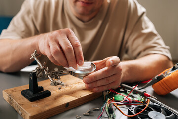 Person focusing on intricate details of circuit board on wooden base with magnifying glass, using helping hands tools and wires during electronic repair or assembly process.
