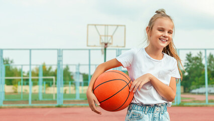 Cheerful child girl basketball player holding game ball standing on the basketball court outdoors