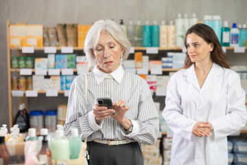 Elderly woman with mobile phone in hands standing in pharmacy and choosing medicines and pills, she flipping through shopping list on mobile phone. Pharmacist advises a client in a pharmacy