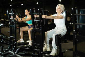 In gym, senior woman uses leg abduction machine to strengthen muscles. Series of repetitions to consolidate result. Perseverance and persistence in achieving fitness goal.