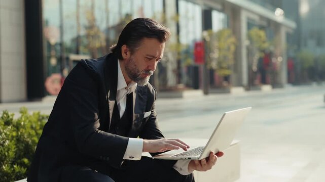 Portrait of elderly corporate CEO man types on laptop keyboard as sits on parapet in downtown. Mature businessman writes email on notebook computer working outside office building