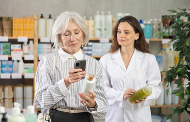 Mature woman in pharmacy scans QR code on shampoo product, checks certificate on manufacturers website and consulting with woman pharmacist about using cosmetic care product