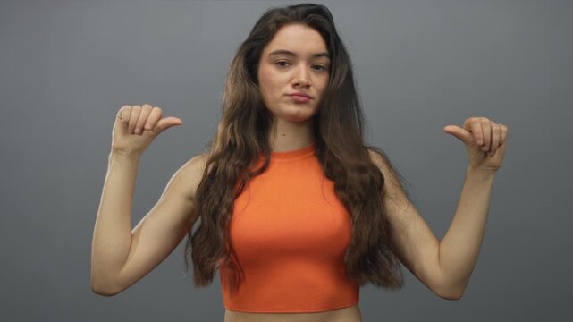 Hispanic young woman in orange crop top points thumbs to chest in grey studio; confidence self esteem empowerment.