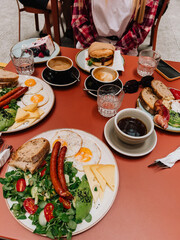 Table filled with brunch dishes like sausages, eggs, cheese, salad, burger, cheesecake, surrounded by coffee cups, capturing friends enjoying meal together in cafe. Friends sharing meal at cafe table