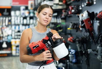 Fototapeta premium Beautiful young girl planning home renovation and choosing paint sprayer in hardware store