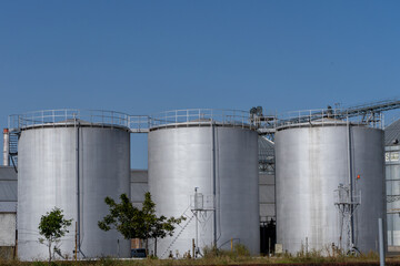 Three large silver grain silos stand tall under a clear blue sky, part of an extensive agricultural storage complex.