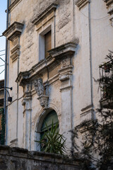 baroque architecture detail of an ancient, weathered stone building facade in southern italy, featuring ornate carvings, an arched window, and a plant peeking through, under warm daylight