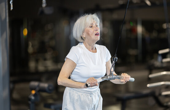 Fit grey-haired senior woman performing triceps pulldown on cable machine in well equipped gym, showing confidence, strength, and care for body at any age