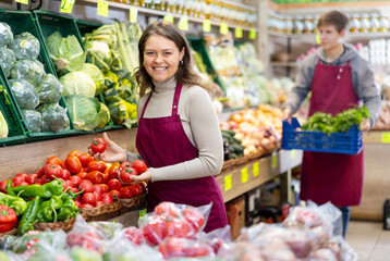 Young woman seller in uniform lays out tomatoes on counter in grocery store