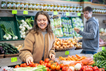 Young woman shopper in casual clothes chooses tomatoes in vegetable shop