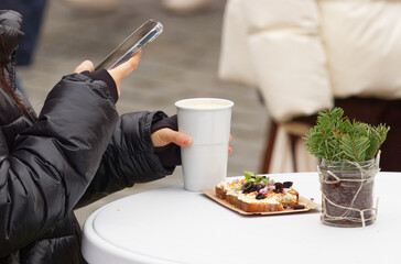 Visitor at Prague Naplavka winter market holding coffee and phone, taking a photo of open-face sandwich on table with small festive decoration in cold advent season.