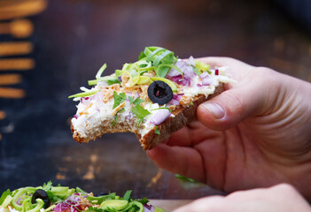 Hand holding bitten open-face sandwich with rich spread and colorful veggies at Prague Naplavka market, capturing the moment of eating in cool outdoor weather.