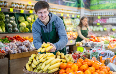Young salesman in apron lays out bananas on counter in vegetable shop..