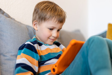 Portrait of cute adorable blond little kid boy enjoy sitting on home sofa surf web play game or watching TV show movies using tablet. Children smart technology usage concept. Child gadget addiction