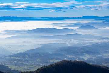 Obraz premium Wide panoramic view across valleys filled with dense fog, showcasing layered mountain ridges and the hill town of Kamnik in the foreground