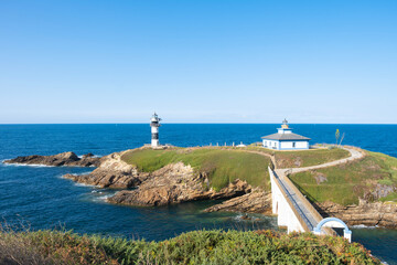 Scenic landscape of Isla de Pancha in Ribadeo, Spain, featuring two historic lighthouses on a rocky island connected by a bridge under a clear blue sky