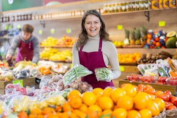 Positive young female seller holding green beans in polybag standing by counter in vegetable market