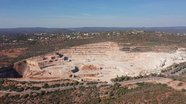 open pit quarry panorama showing expansive benches, heavy machinery tracks, haul roads circling pit, processing area and distant ridgelines, bright clear sky, operational logistics