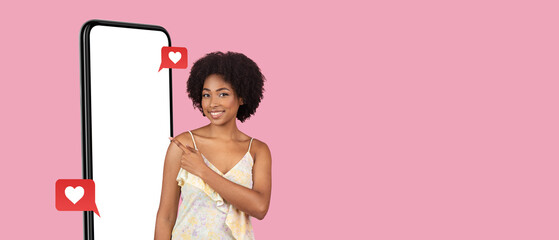 A young woman stands next to a large smartphone with a blank screen, smiling and pointing towards social media like icons on a pink background, showcasing digital engagement.