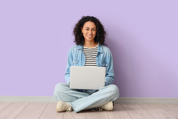 Young African-American woman working with laptop near lilac wall