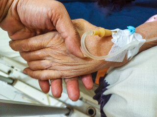 Close-up of a wrinkled hand gently holding the hand of a patient with an IV drip (cannula) on a hospital bed. Concept for senior care, compassion, health insurance, and medical support.