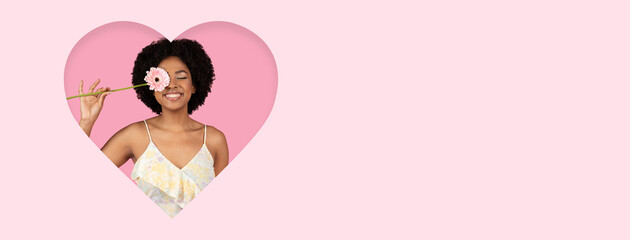 A joyful woman with curly hair smiles while holding a flower in front of her face. She stands against a soft pink heart-shaped backdrop, radiating positive energy and happiness.
