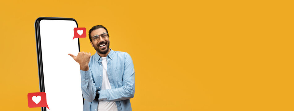 A cheerful man stands beside a blank smartphone on a vibrant yellow background, making a thumbs-up gesture. Notification icons with hearts float around him, adding a lively touch. - Powered by Adobe