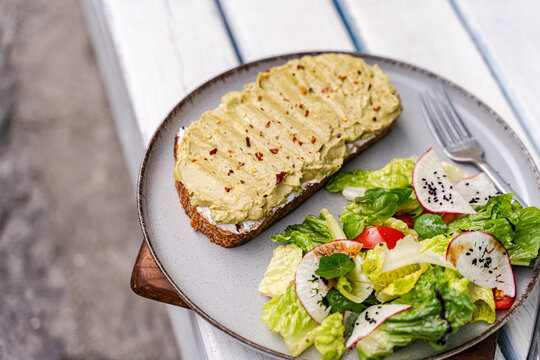 Overhead view of a plate of Avocado toast with cream cheese, chilli flakes and a mixed salad on a garden table