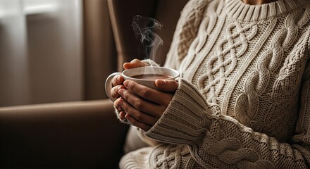 A person in a cozy cable-knit sweater holding a steaming hot mug of coffee or tea indoors.
