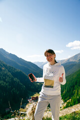 Smiling travel blogger woman showing thumbs up on Transfagarasan Highway in Romania, enjoying sunny outdoor scenery with deep forested valleys and clear sky. Ideal for lifestyle travel content