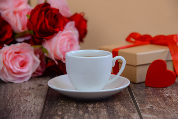 A cup of hot coffee with roses and a gift with a red ribbon on the table