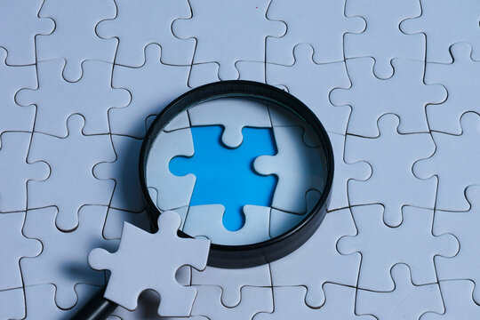 Magnifying glass focusing on the missing gap of a white puzzle, with the missing piece placed on the magnifying glass, representing problem solving, gap analysis, and root cause analysis