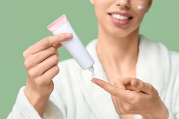 Beautiful young woman in robe with tube of face cream on green background