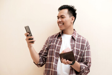 Southeast Asian man wearing flannel shirt standing and smiling expression happy with holding leather wallet and mobile phone, on white isolated background.