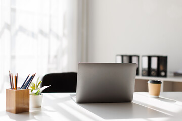 Laptop view of comfortable office desk with laptop, mug, tree pot, notebooks and copy space on white table in glass partition office
