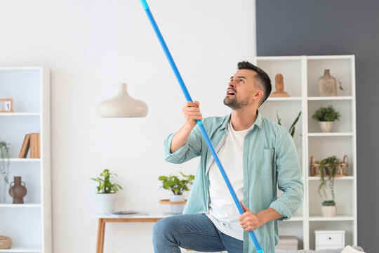 Young man banging ceiling with mop because of loud neighbours at home