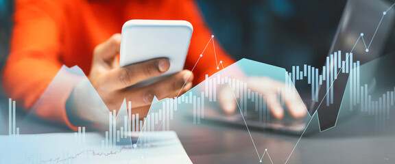 An Indian entrepreneur is engaged in business activities in a dark office space, using a smartphone for mobile applications while analyzing data on his laptop. He examines graphs and trends.