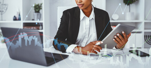 A successful black businesswoman is working diligently in a modern office. She uses a digital tablet and laptop to focus on her tasks.