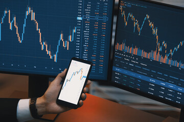 Cropped hand businessman holding smartphone showing dynamic stock market, comparing with financial data report investment graph on two computer monitor screen online at neon light at night. Infobahn.
