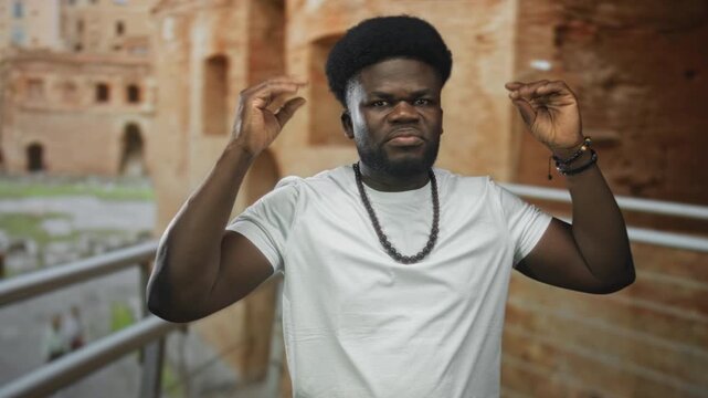 Man with hands raised wearing white tshirt and beaded necklace on roman ruins building balcony railing, gesturing emphatically; determination resilience.