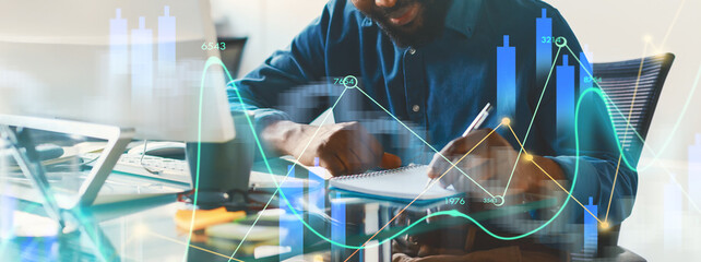 A person is seated at a desk actively taking notes while reviewing business graphs and charts on a computer screen. The setting is a modern office filled with data visualizations.