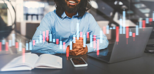 A young Indian professional works at his desk in a modern office, using his laptop for a virtual meeting. Charts displaying business data are visible, highlighting an active work environment.
